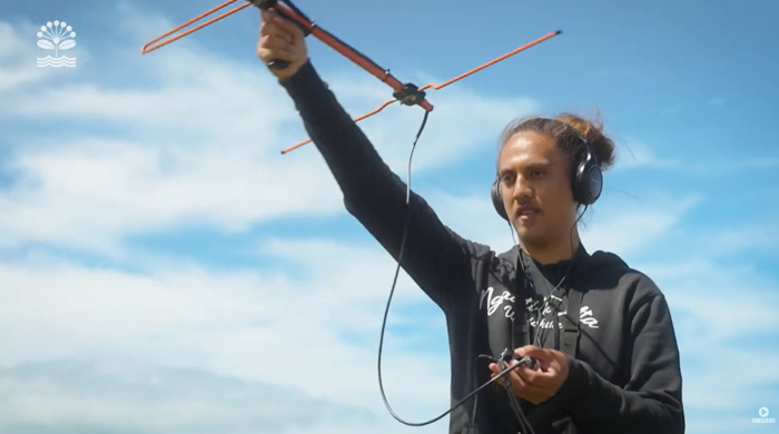 A Māori man stands holding a metal antennae into the air. He wears headphones that are attached to the device.